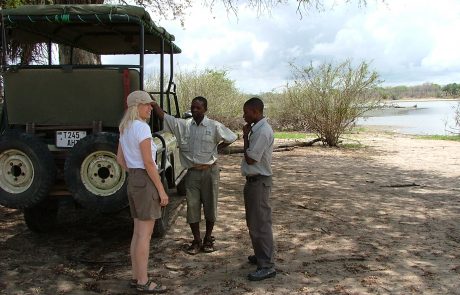 Frau in Shorts & weißem T-Shirt steht mit zwei Guides an einem Safari Fahrzeug im Schatten