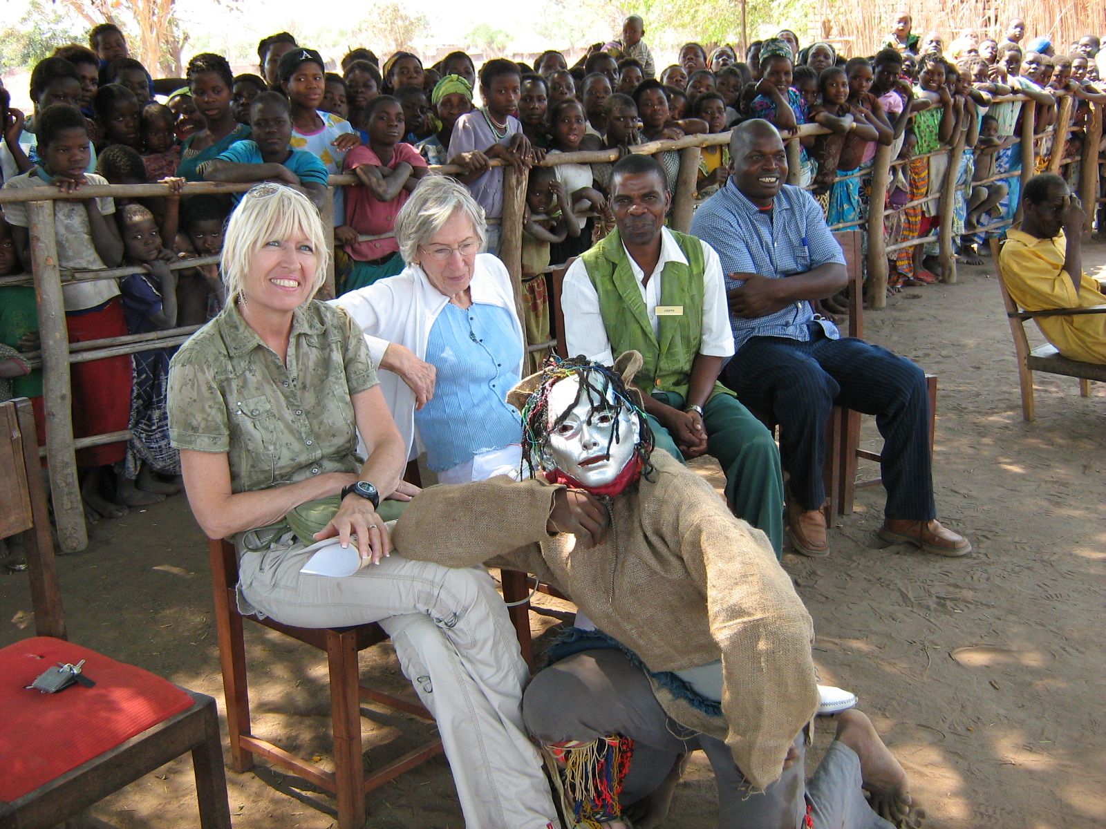 Zwei Frauen sitzen lachend auf einem Stuhl. Ein Mann in Maske kniet vor ihnen. Hinter einem Zaun stehen zahlreiche Dorfbewohner