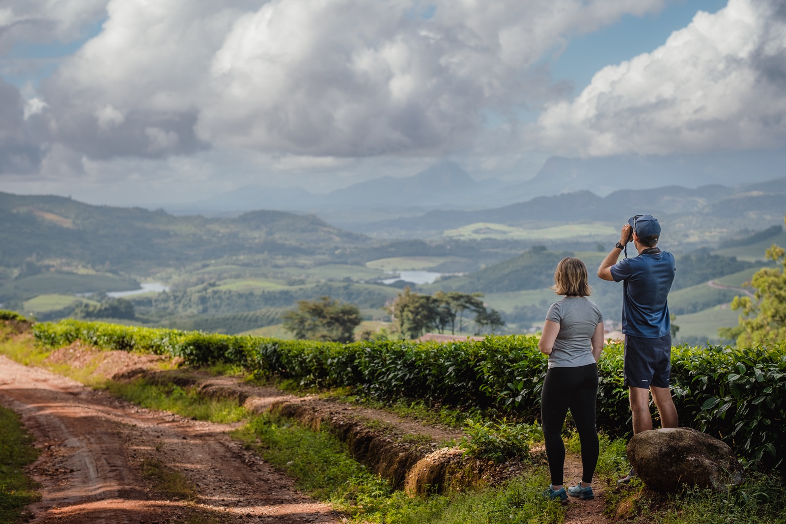 Ein Mann steht am Rand einer roten Sandpiste und blickt durch sein Fernglas in die Ebene, neben ihm eine Frau