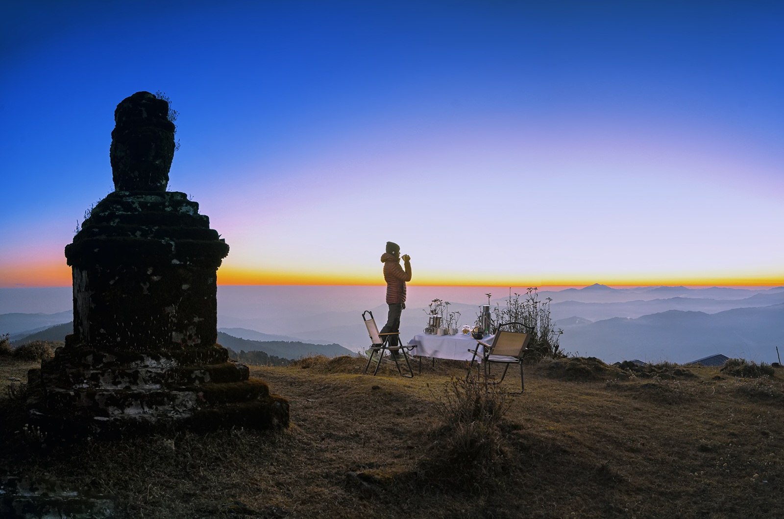 Frau mit dicker Jacke & Mütze hält eine Tasse u. blickt auf die Berge in der Tiefe. Am Horizont Sonnenaufgang