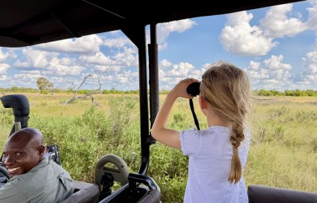 Mädchen steht in einem offenen Safarifahrzeug und blickt durch ein Fernglas in die Landschaft